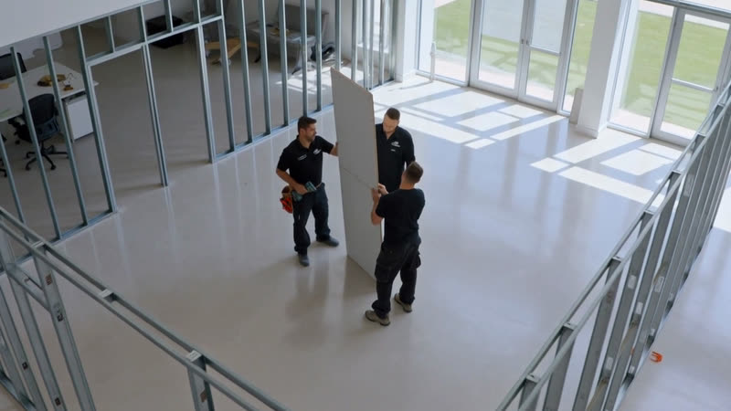 Two installers carrying a plasterboard sheet between steel stud partition frames